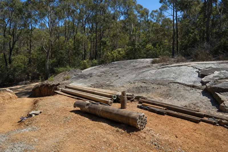 Drill string and a pneumatic down-hole hammer bit laid out on orange-red lateritic gravel beside exposed granite, jarrah forest behind — water bore drilling & installation, Perth. Illustrative example.