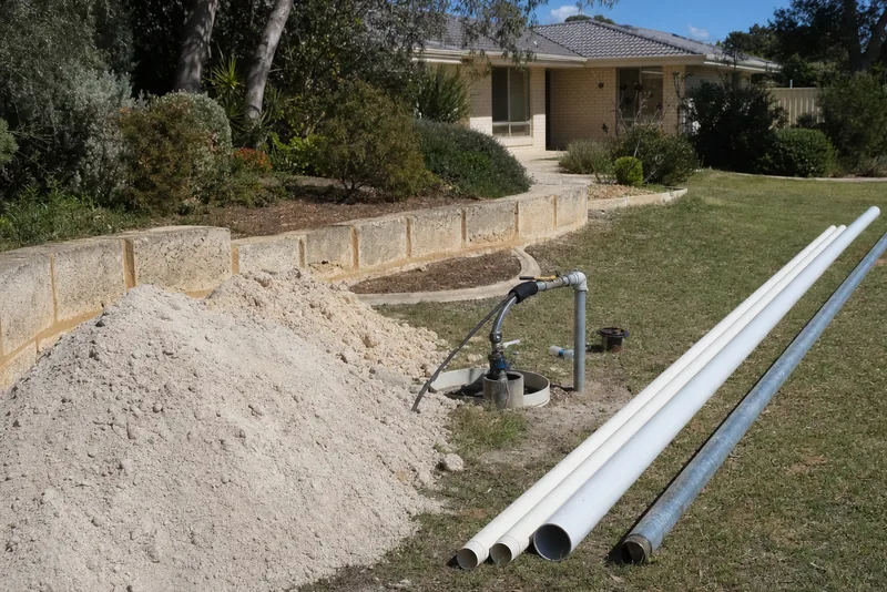 Lengths of PVC bore casing and galvanised riser laid along a lawn beside an open bore head, pale sand spoil and a cut-limestone wall — bore repairs & pump repairs, Perth. Illustrative example.