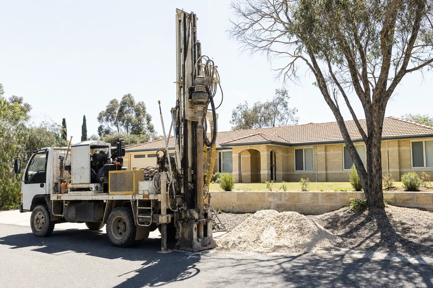 A truck-mounted water bore drilling rig set up on the driveway of a single-storey Perth home, mast raised, pale sand heaped beside the borehole — illustrative example